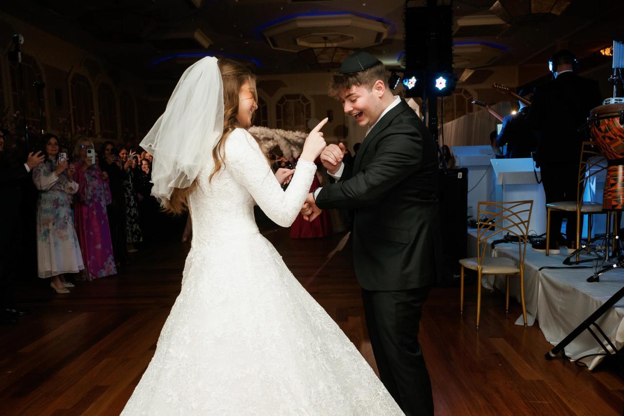 Bride and groom dancing together on the reception floor, smiling and pointing, with guests and a stage in the background under blue lighting. Wedding photography at The Palace, Brooklyn, NY.