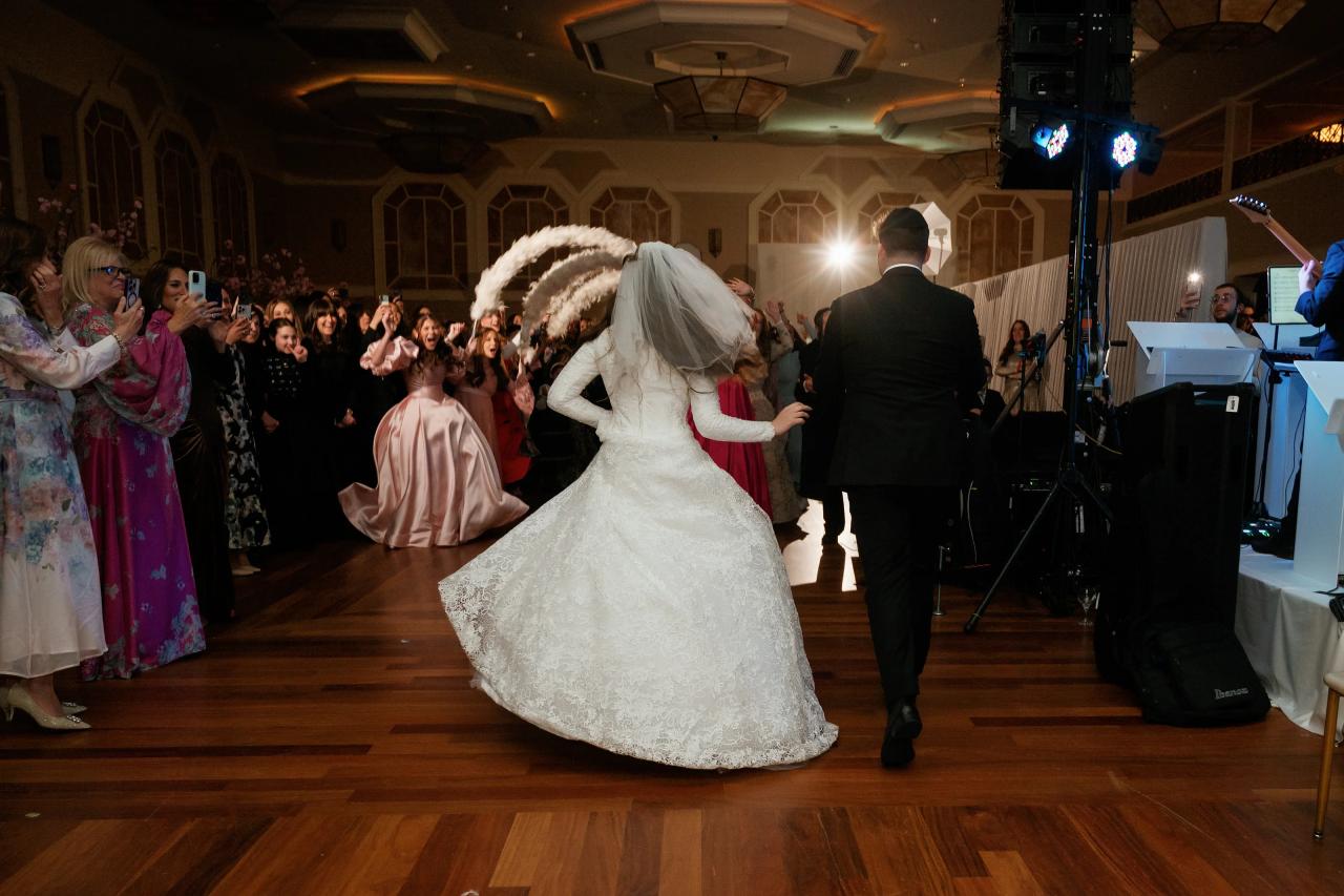 Bride in a white wedding gown twirling on a ballroom dance floor while the groom faces away toward the DJ area, with guests cheering in the background under warm stage lights. Wedding photography at The Palace, Brooklyn, NY.