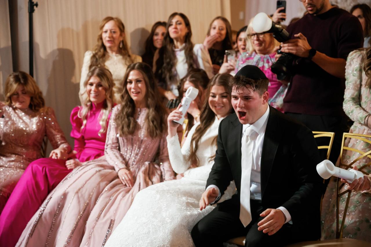 Laughing groom leaning forward holding a party horn while the bride smiles at a lively wedding reception, with guests cheering in the background. Wedding photography at The Palace, Brooklyn, NY.