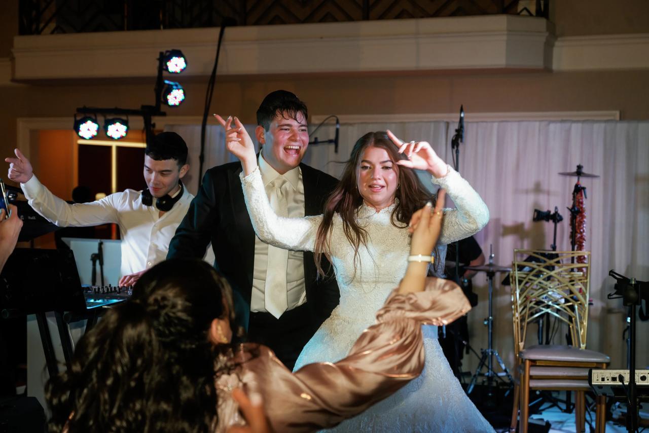 Frum wedding dance-floor moment: bride and groom smiling with both hands raised while dancing in an event hall with stage lights and a DJ in the background. Wedding photography at The Palace, Brooklyn, NY.