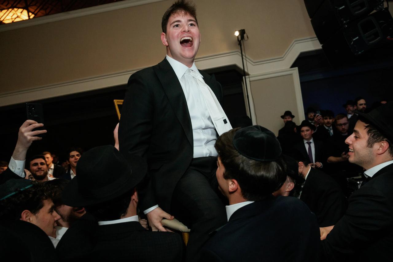Groom laughing with mouth open during a lively dance celebration, surrounded by guests in a reception hall with stage lights. Wedding photography at The Palace, Brooklyn, NY.