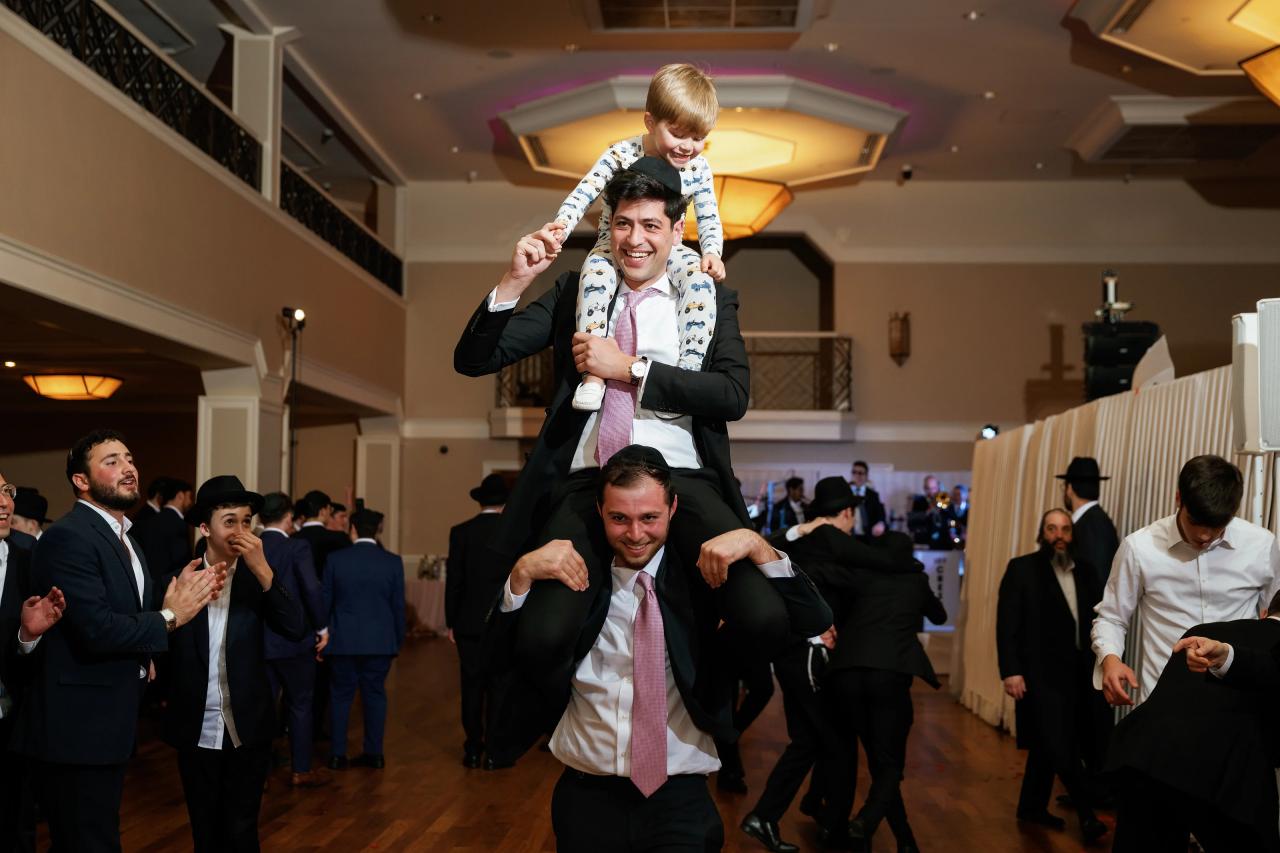 A groom smiling while lifting a small child on his shoulders during a lively reception dance, with guests clapping in the background. Wedding photography at The Palace, Brooklyn, NY.