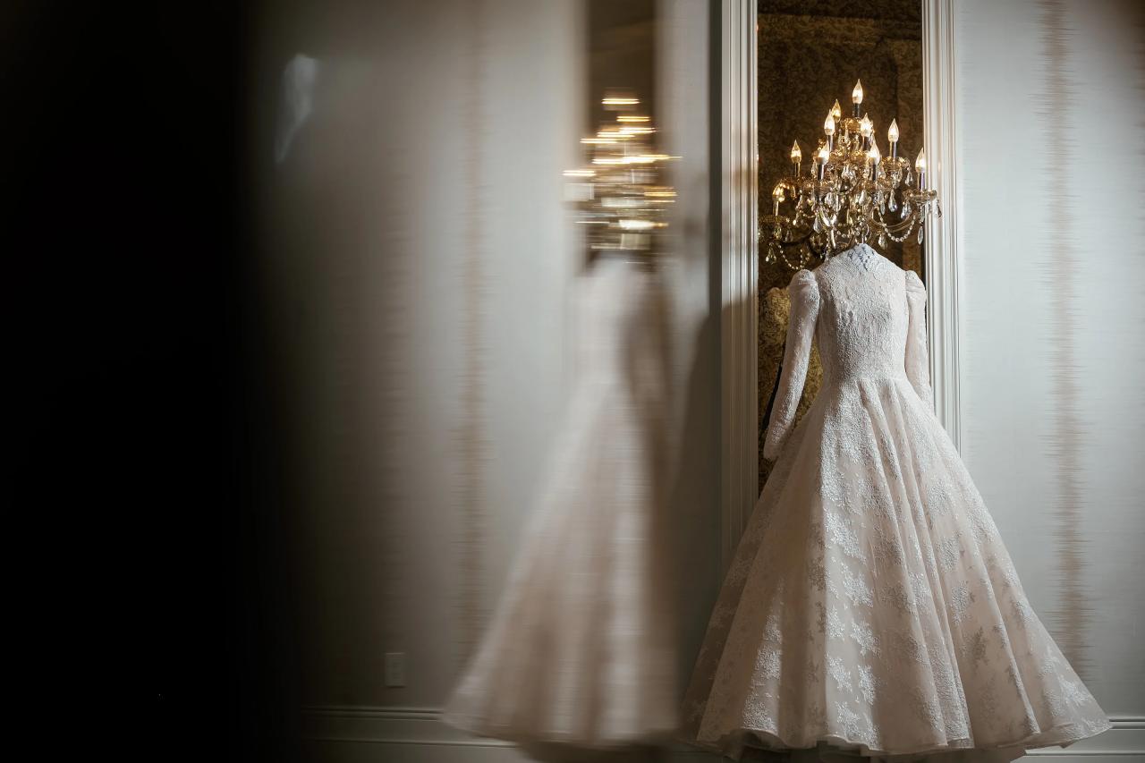 A lace wedding gown hangs beside a crystal wall light, reflected softly in the foreground. From this Lake Terrace wedding. Wedding photography.