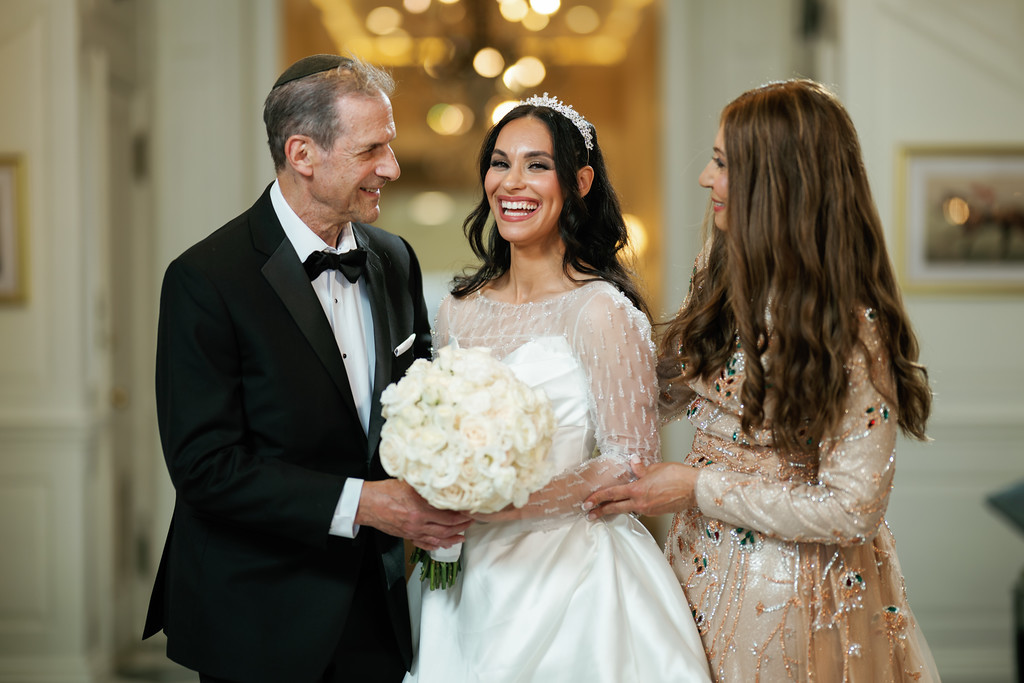 Smiling bride in a white wedding gown and tiara holding a bouquet as her father in a black tuxedo stands beside her and a woman in a beaded dress looks on during the event.