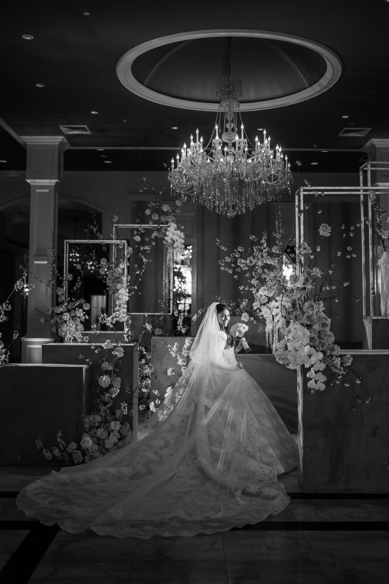 Black-and-white full-length bridal portrait beneath a chandelier, surrounded by tall floral displays. From this Lake Terrace wedding. Wedding photography.