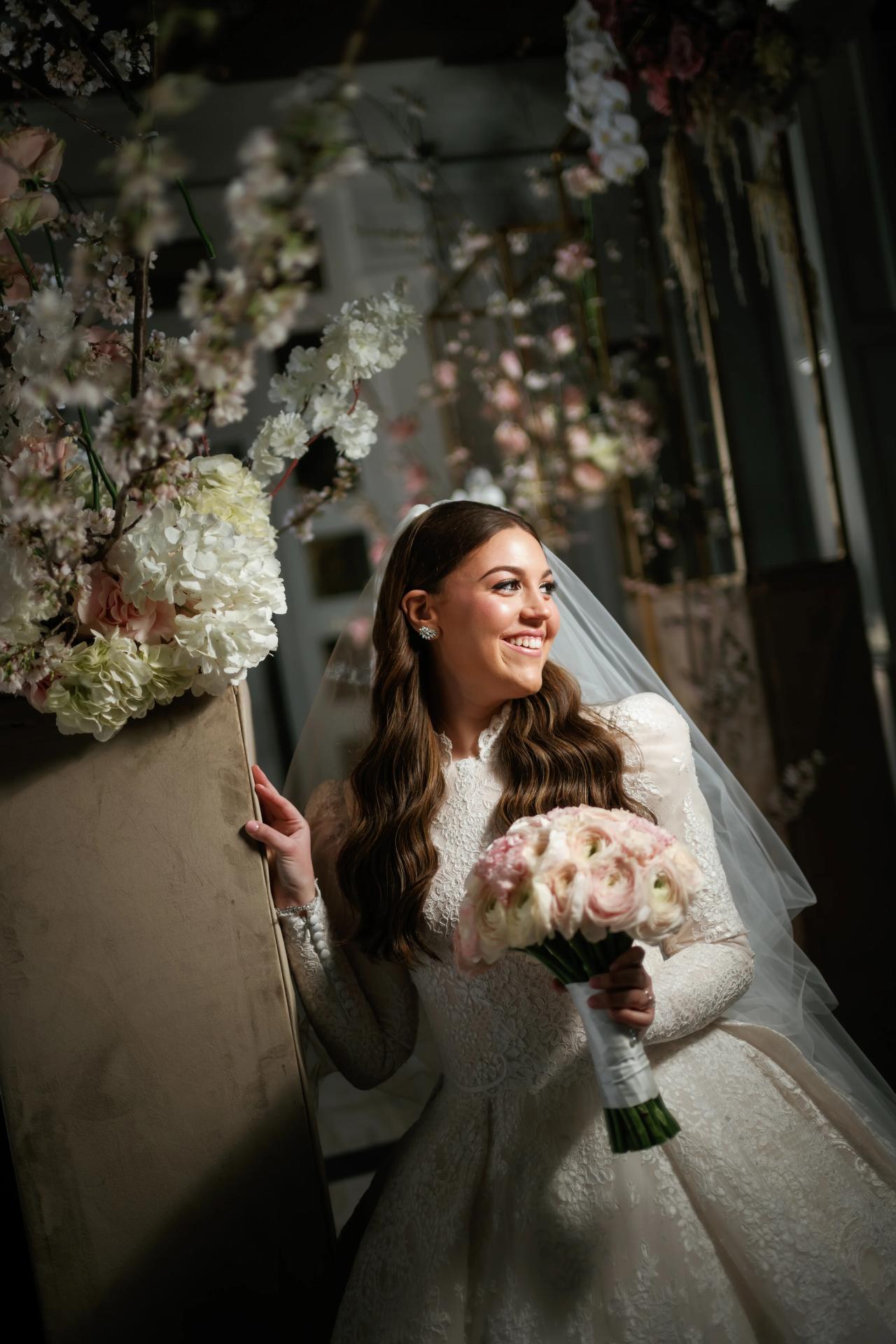 The bride smiles beside tall white and blush flowers while holding her rose bouquet. From this Lake Terrace wedding. Wedding photography.