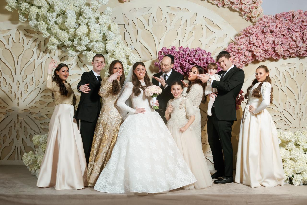 The bride poses with a lively group of adults and children against sweeping white and pink flowers. From this Lake Terrace wedding. Wedding photography.
