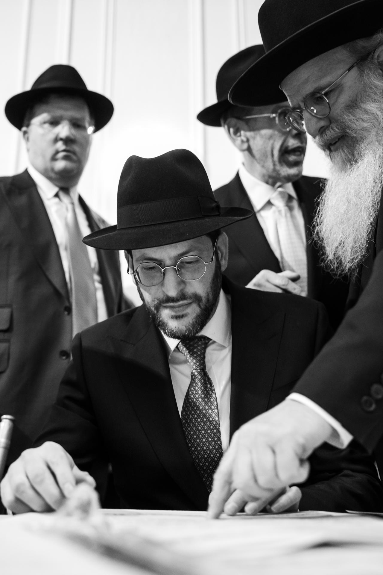 Black-and-white view of men gathered closely around a document during the wedding preparations. From this Lake Terrace wedding. Wedding photography.