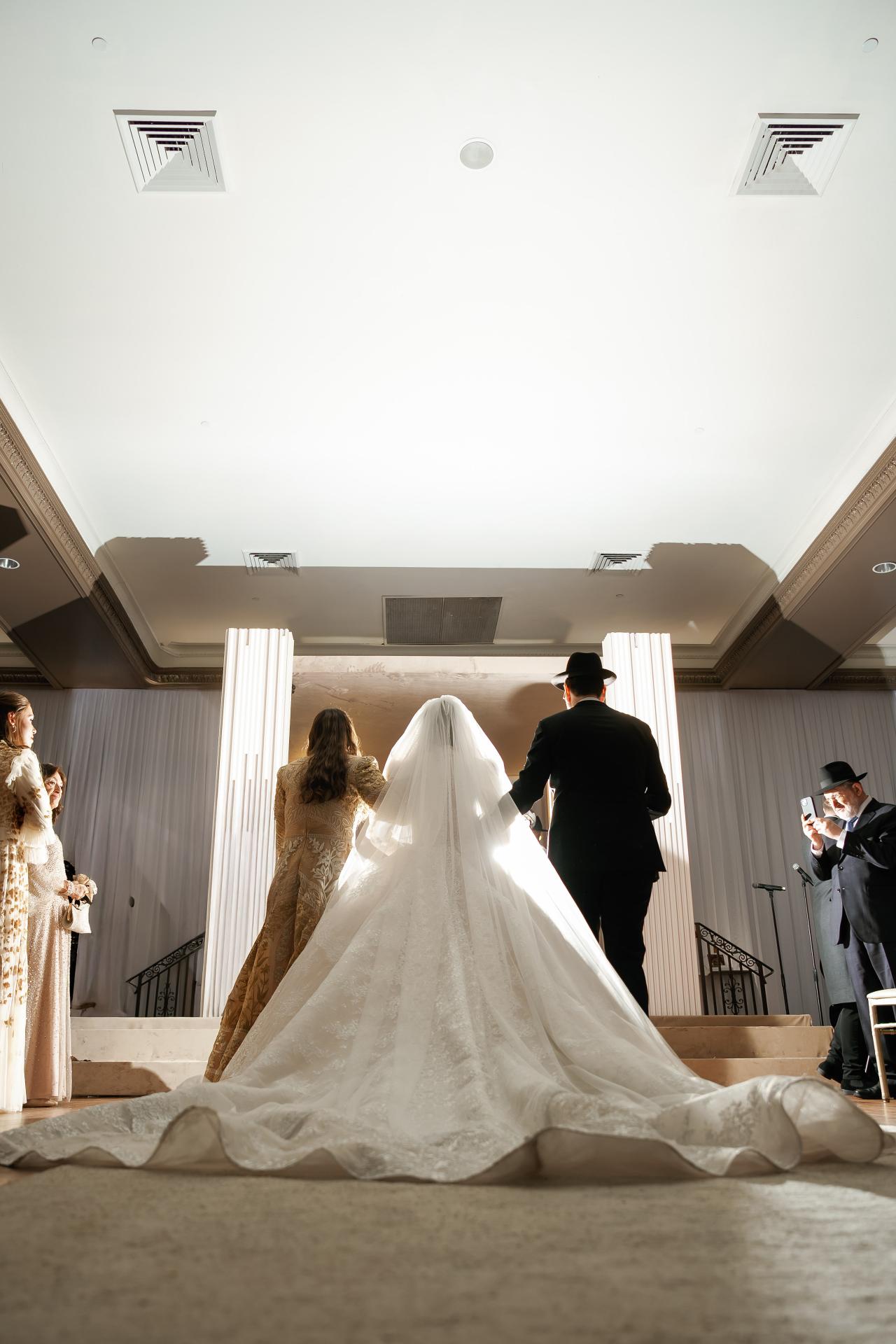 Low-angle view of the bride’s lace train as she approaches the white-draped chuppah. Wedding photography at Lake Terrace, Lakewood, NJ.