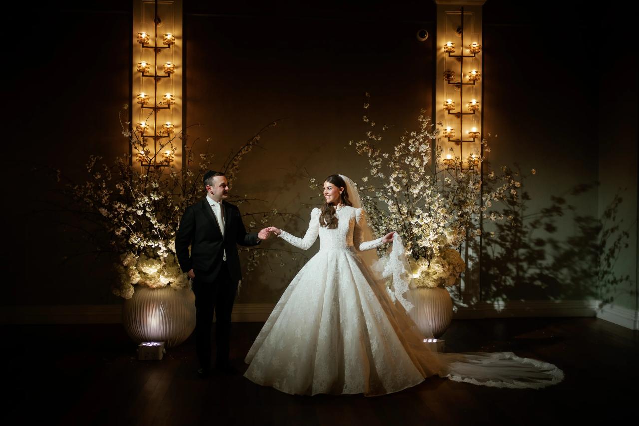 Bride and groom holding hands in warm golden light beside white blossoms. Wedding photography at Lake Terrace, Lakewood, NJ.
