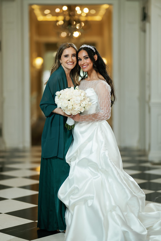 Two women smiling together in an elegant hallway; one holds a white rose bouquet while the other stands in a white bridal-style gown and veil.