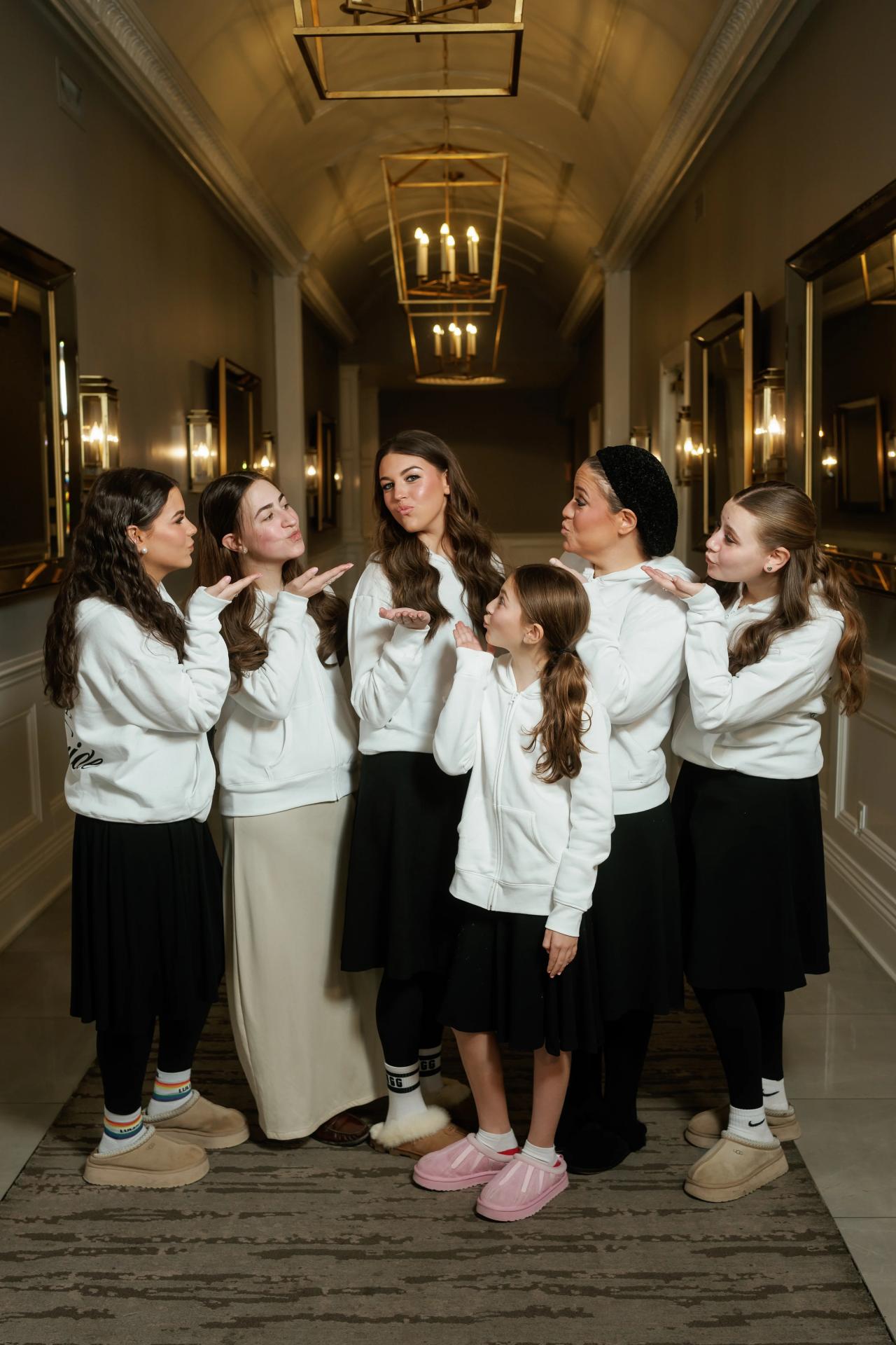 Group of young girls in white hoodies and dark skirts posing in a hotel hallway, making finger heart gestures under warm chandelier light. Wedding photography at Lake Terrace, Lakewood, NJ.