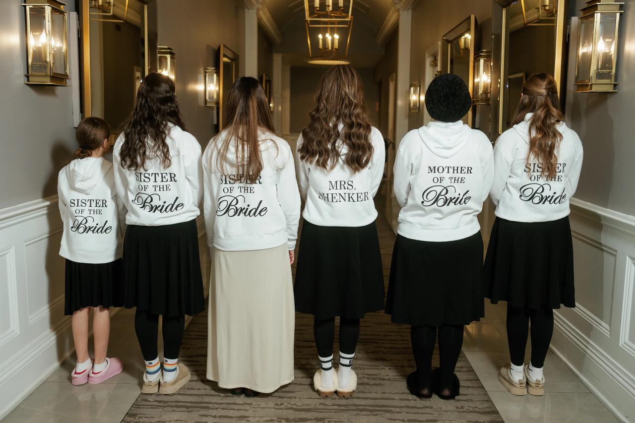 Six people in coordinated bride-themed hoodies pose along a warmly lit hallway, facing away from the camera. Wedding photography at Lake Terrace, Lakewood, NJ.