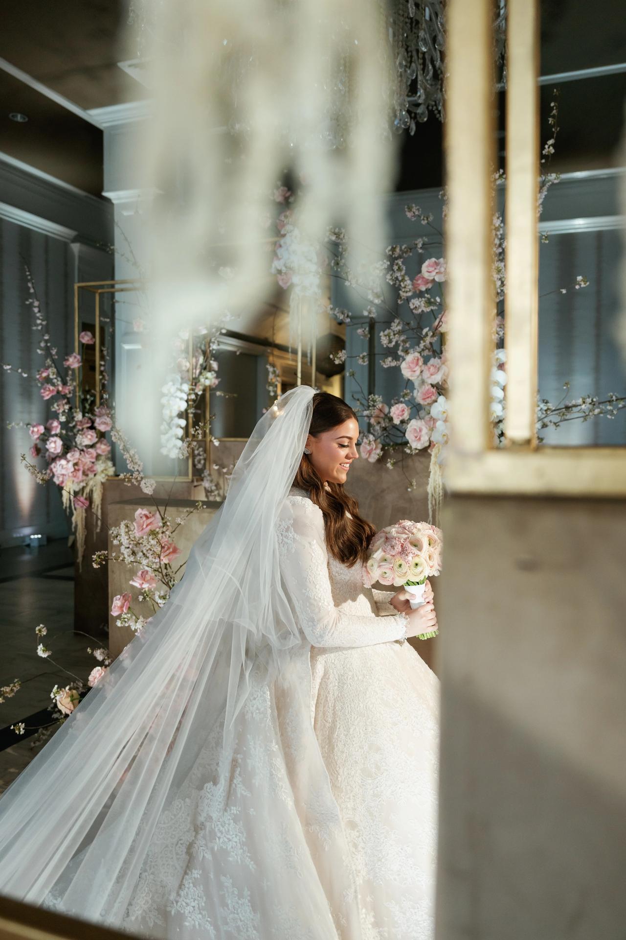 Framed profile portrait of a bride in a white gown holding a bouquet, her veil flowing through warm candle and floral corridor light with soft lens flare. Wedding photography at Lake Terrace, Lakewood, NJ.