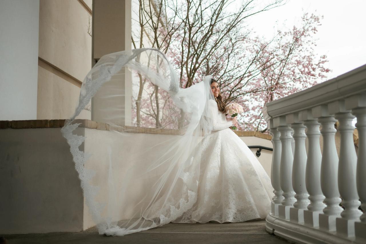 Bride in a flowing white gown and veil smiling on a balcony with soft daylight and pink blossoms in the background, holding a bouquet. Wedding photography at Lake Terrace, Lakewood, NJ.