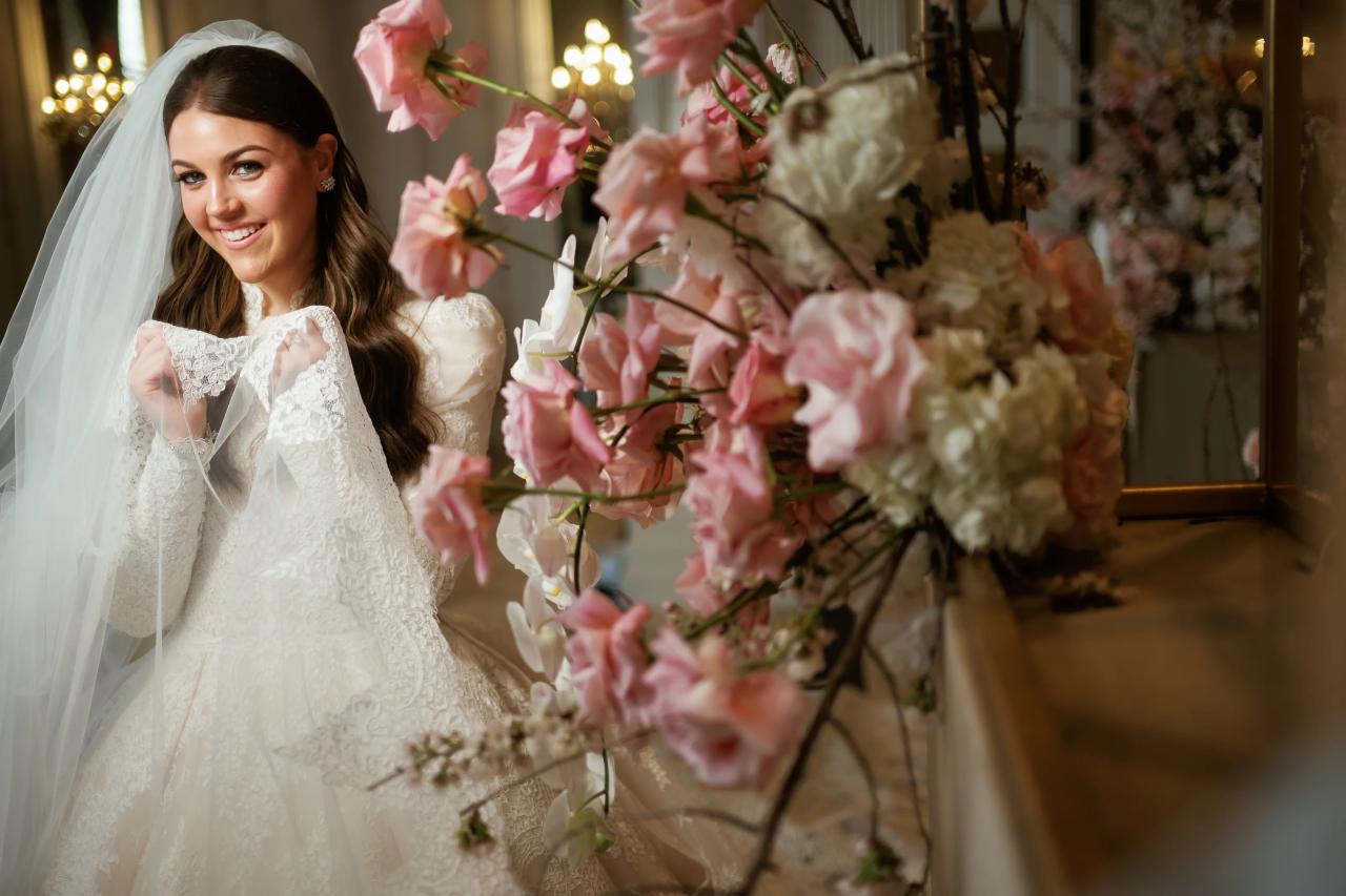 Smiling bride in lace holding her bouquet, framed by pink floral branches with warm chandelier light in the background. Wedding photography at Lake Terrace, Lakewood, NJ.