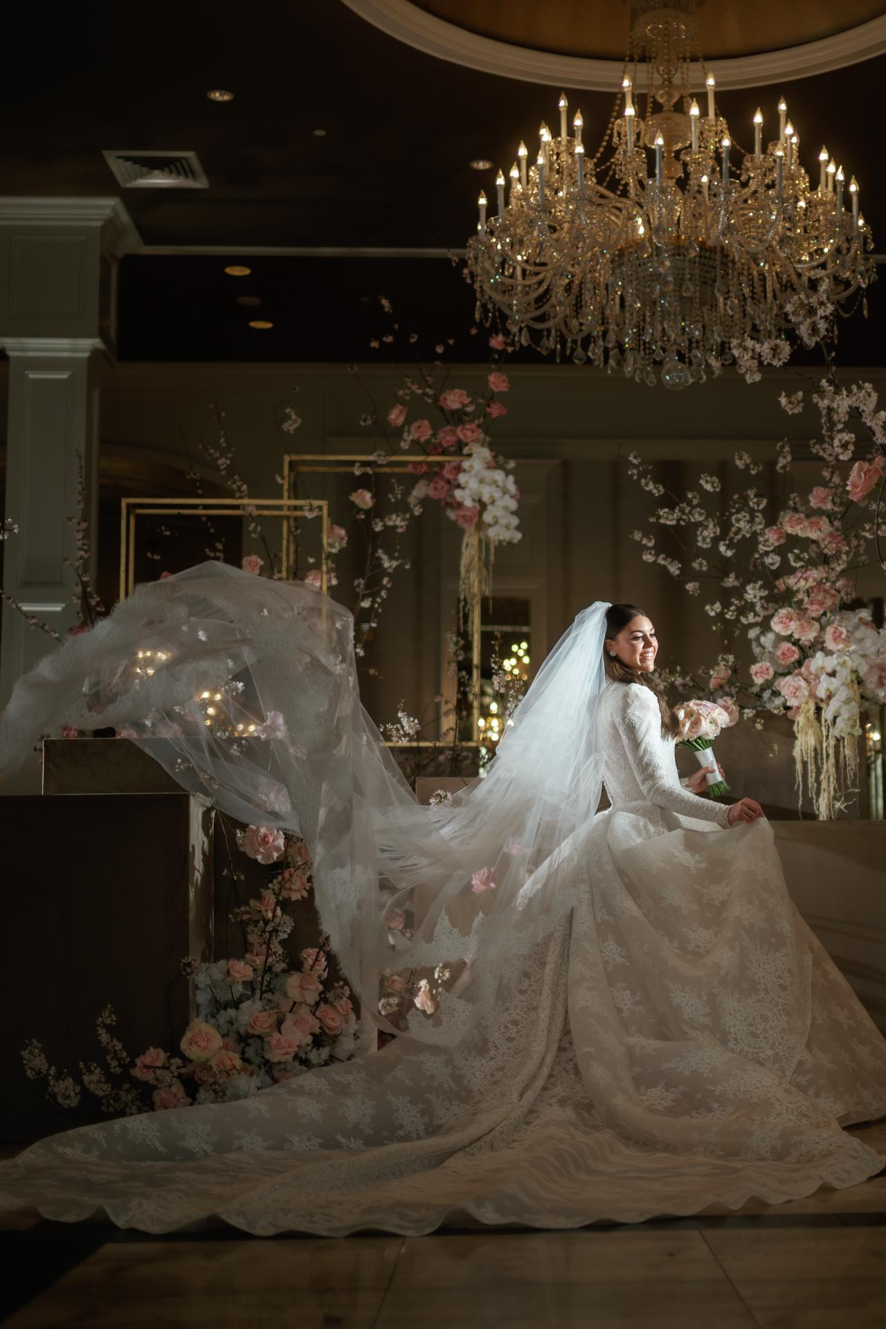 Bride in a white veil and gown walking forward outdoors-illusionously indoors, holding an open umbrella, smiling in front of a floral ceremony backdrop under a chandelier. Wedding photography at Lake Terrace, Lakewood, NJ.