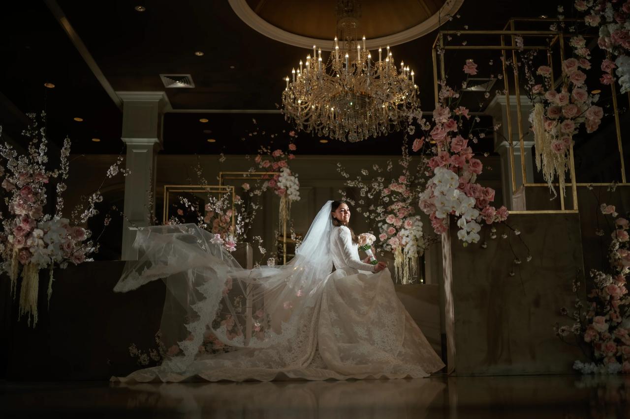 A bride in a flowing white gown and veil moves through a floral ceremony setup beneath a chandelier, with pink and white flowers framing the scene and warm lights in the background. Wedding photography at Lake Terrace, Lakewood, NJ.