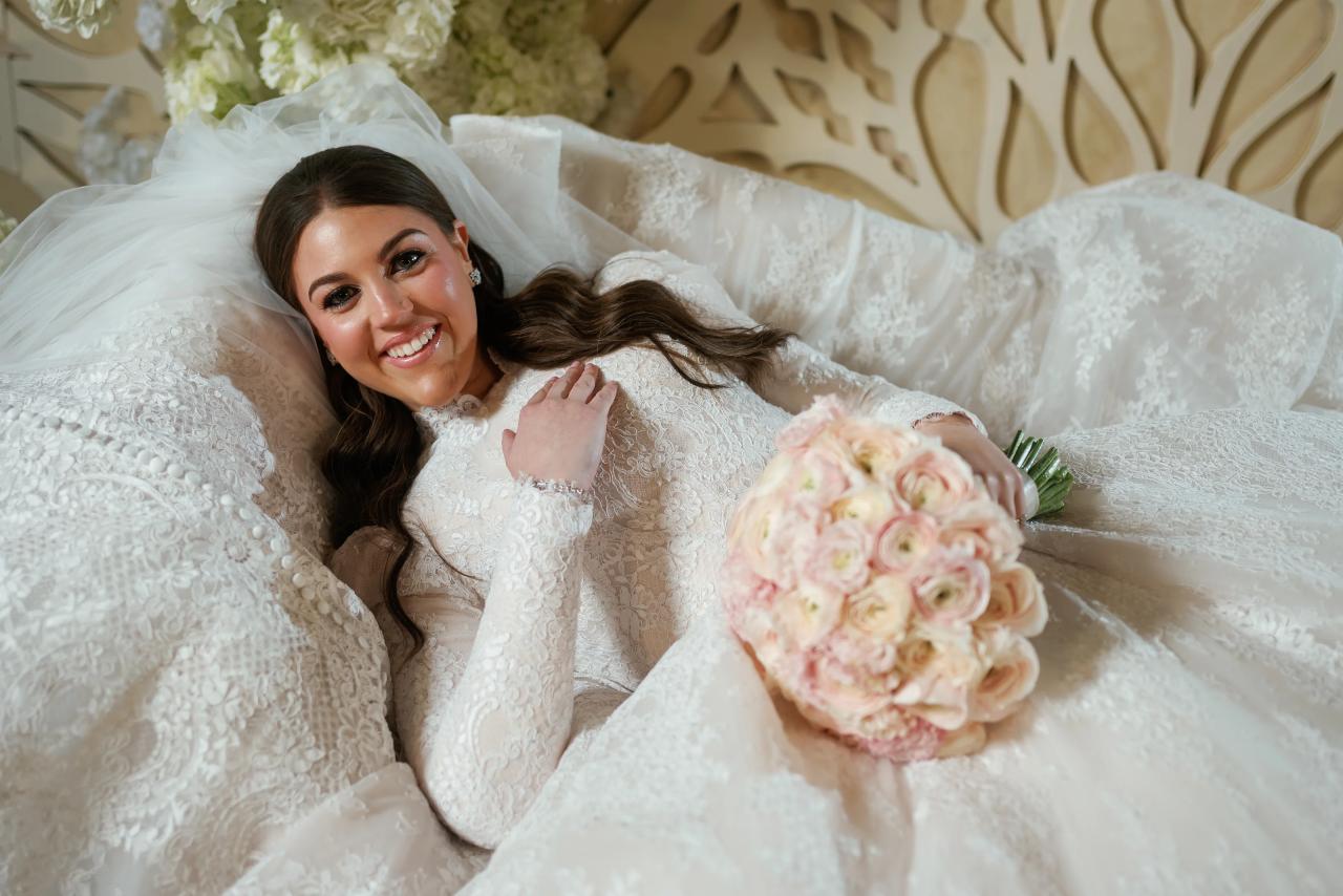 Smiling bride reclining in a white bridal setting with a floral backdrop, veil draped, bouquet in the foreground. Wedding photography at Lake Terrace, Lakewood, NJ.