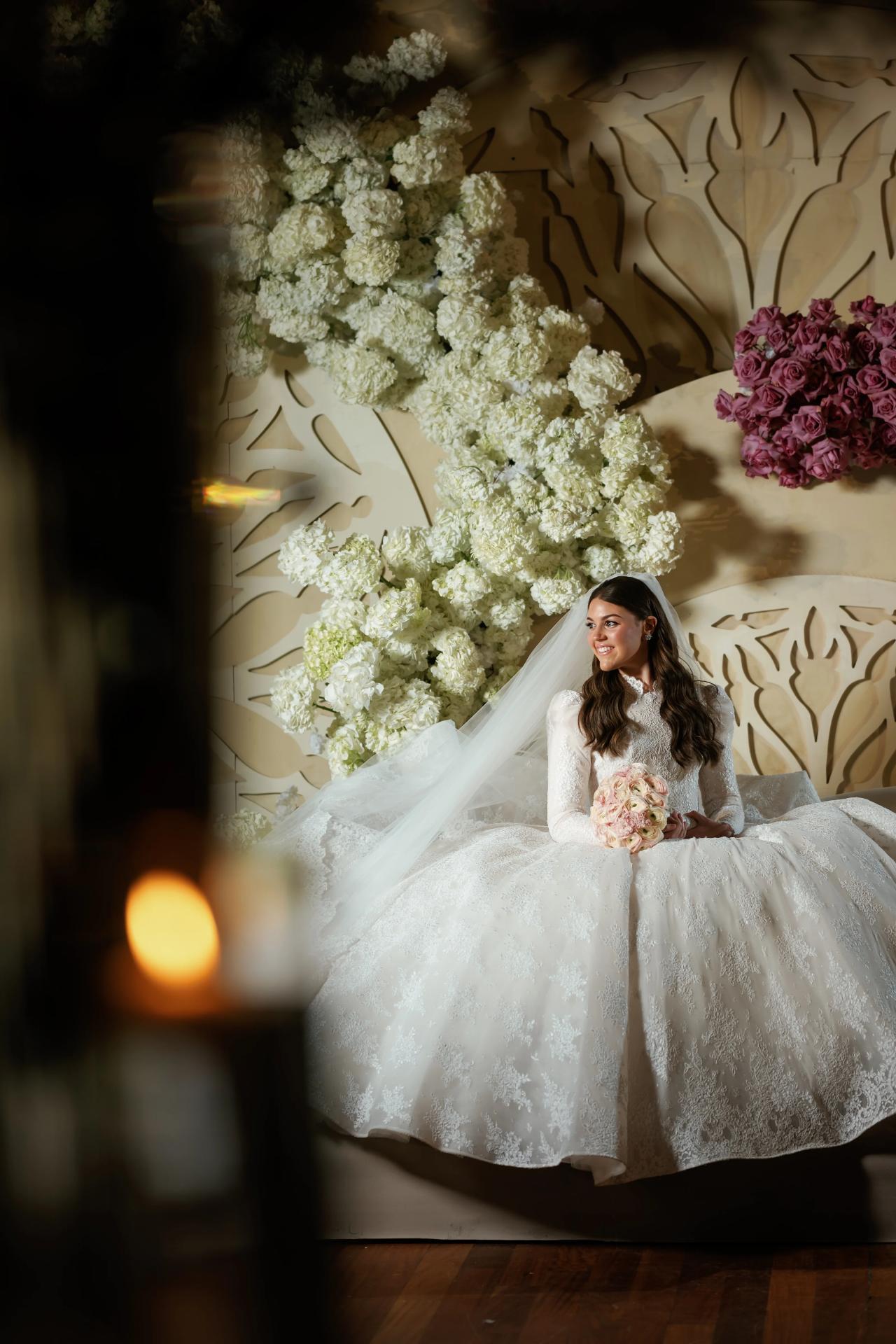Bride in a white gown seated in front of a large floral backdrop, holding a bouquet and smiling in warm, softly lit evening light. Wedding photography at Lake Terrace, Lakewood, NJ.