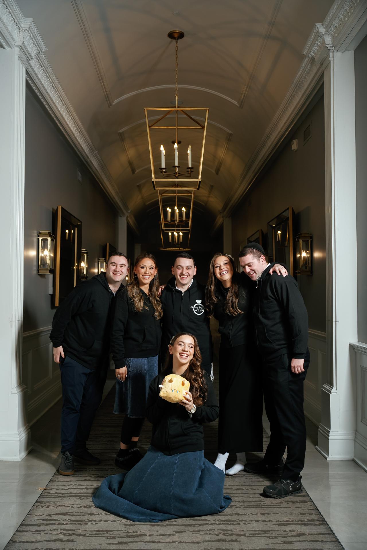 Family posing together down a long hallway in warm light, with a child kneeling in the foreground holding a snack prop. Wedding photography at Lake Terrace, Lakewood, NJ.