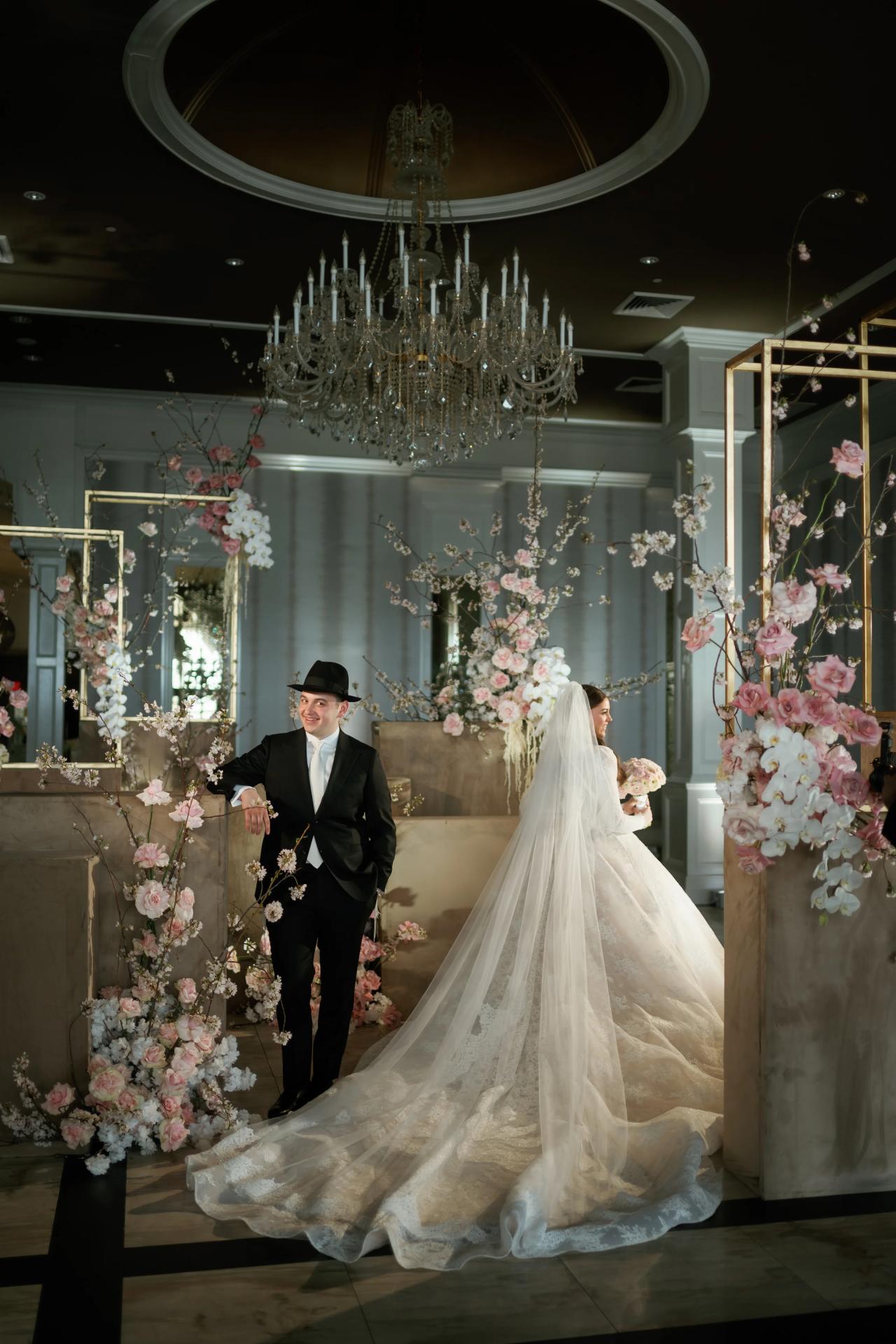 Bride and groom walking hand-in-hand through a floral aisle, with the bride’s long veil flowing under warm chandelier light. Wedding photography at Lake Terrace, Lakewood, NJ.