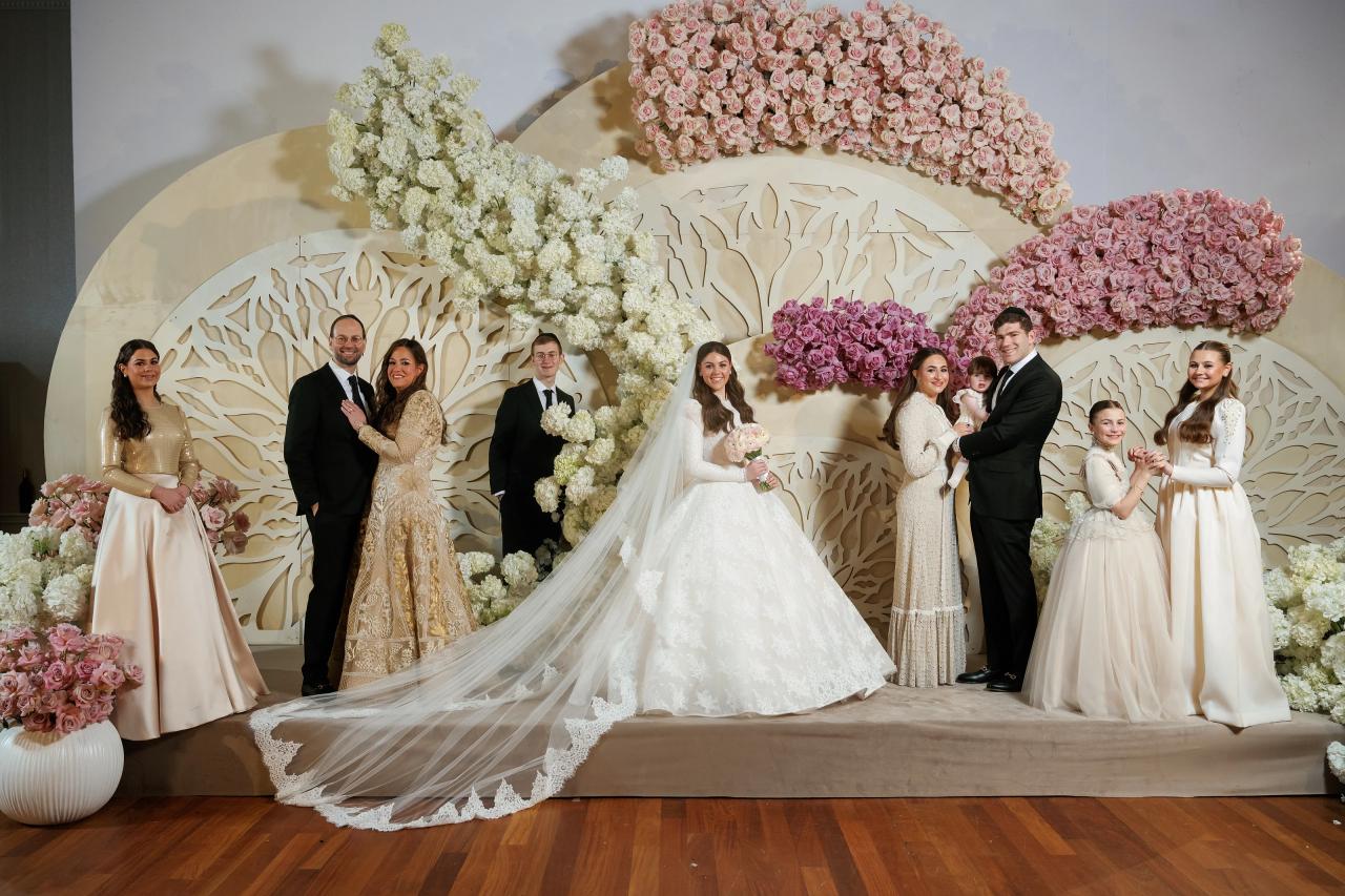 Wedding couple and extended family posing on a floral stage with pastel white and pink arrangements, bride in a flowing gown and veil, groom in a dark suit, surrounded by relatives and children. Wedding photography at Lake Terrace, Lakewood, NJ.