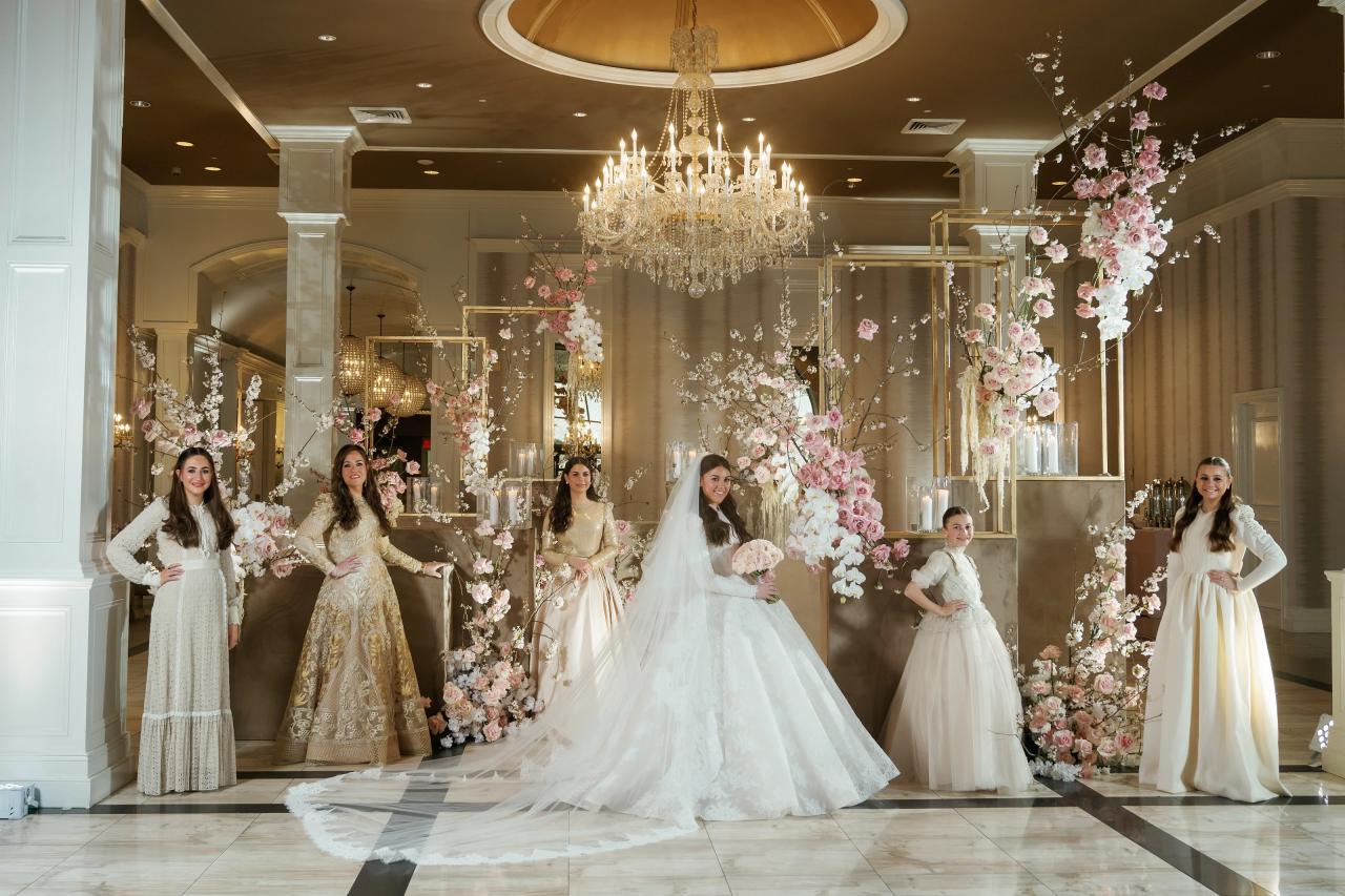 Bride and bridal party walking forward through a floral aisle with chandeliers overhead, petals floating in the warm indoor light. Wedding photography at Lake Terrace, Lakewood, NJ.