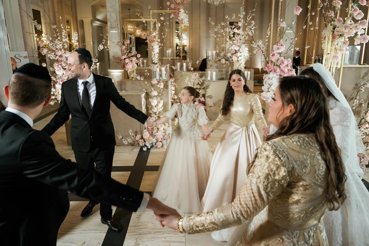 Bride and groom walking through an aisle as guests reach out with open hands, surrounded by pink and white floral arches in a warmly lit ballroom. Wedding photography at Lake Terrace, Lakewood, NJ.