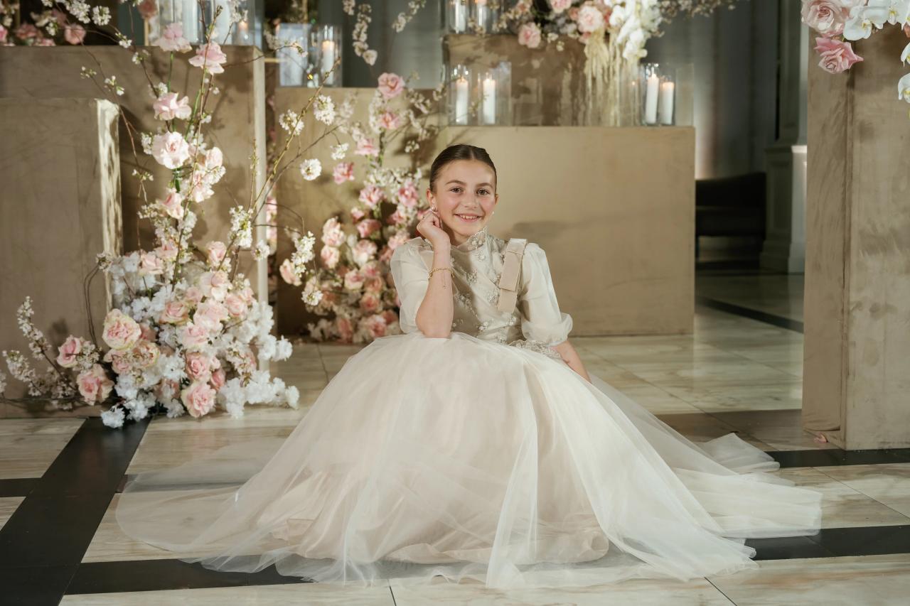 Smiling bat mitzvah girl in an embellished light-colored outfit seated in a formal pose, framed by a pink-and-white floral backdrop in an indoor venue. Wedding photography at Lake Terrace, Lakewood, NJ.
