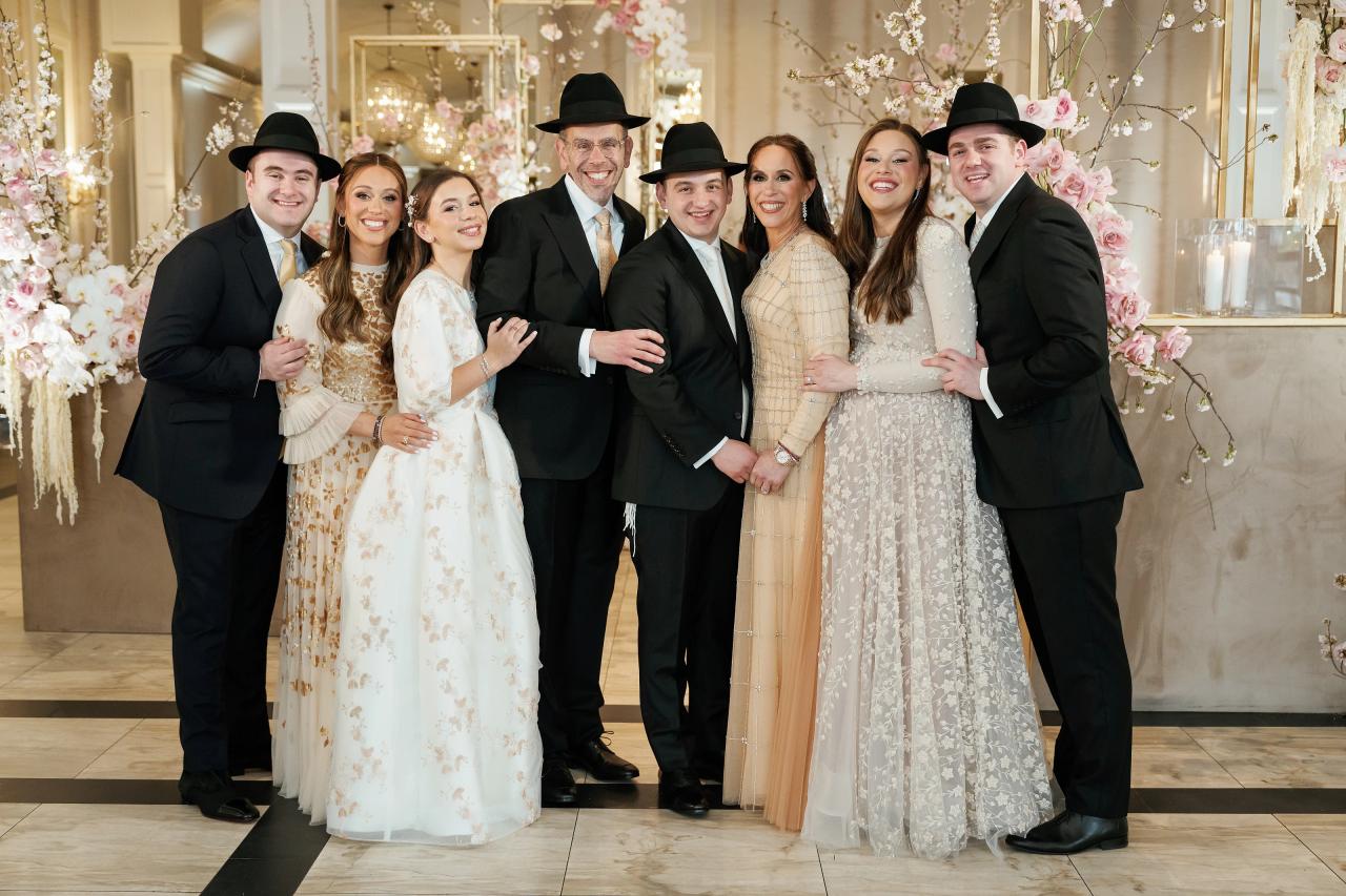 Smiling group of wedding guests posed together indoors, some wearing black hats, in front of floral décor and warm lighting. Wedding photography at Lake Terrace, Lakewood, NJ.
