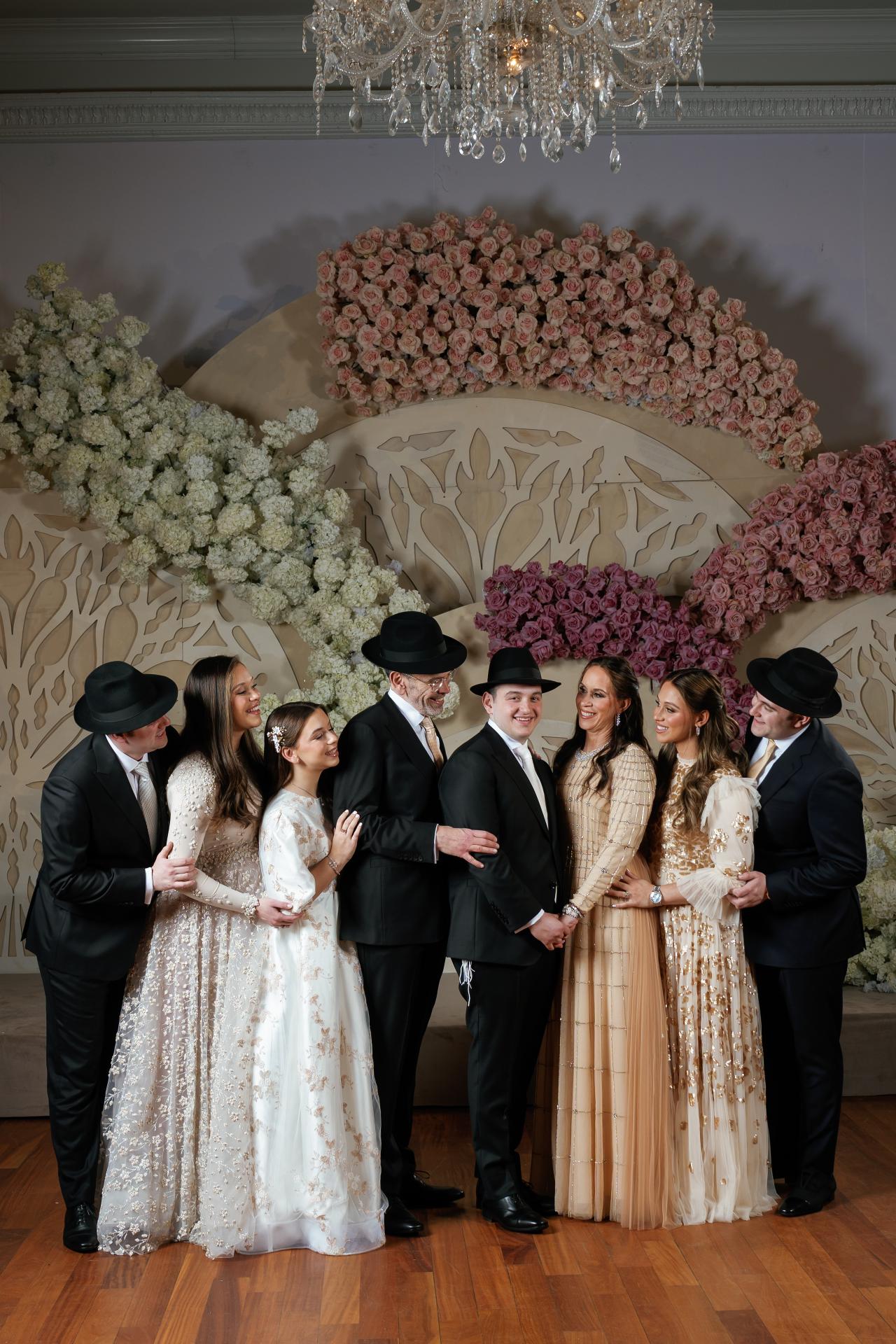 Posed family celebration with the bride and groom in the center in front of a floral rose backdrop, surrounded by guests in formal attire. Wedding photography at Lake Terrace, Lakewood, NJ.