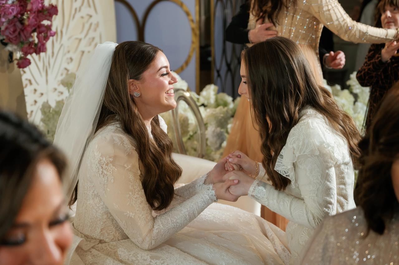Two brides in lace attire and a veil smile while holding hands at a wedding reception, with floral decor and softly blurred guests in the background. Wedding photography at Lake Terrace, Lakewood, NJ.