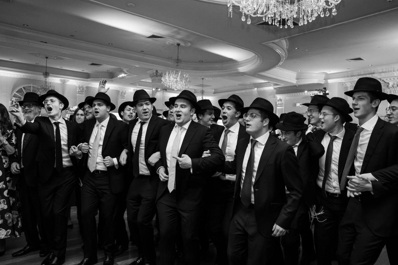 Black-and-white candid of a group of men in dark suits and hats cheering and reacting energetically at a party, with open mouths and raised hands. Wedding photography at Lake Terrace, Lakewood, NJ.