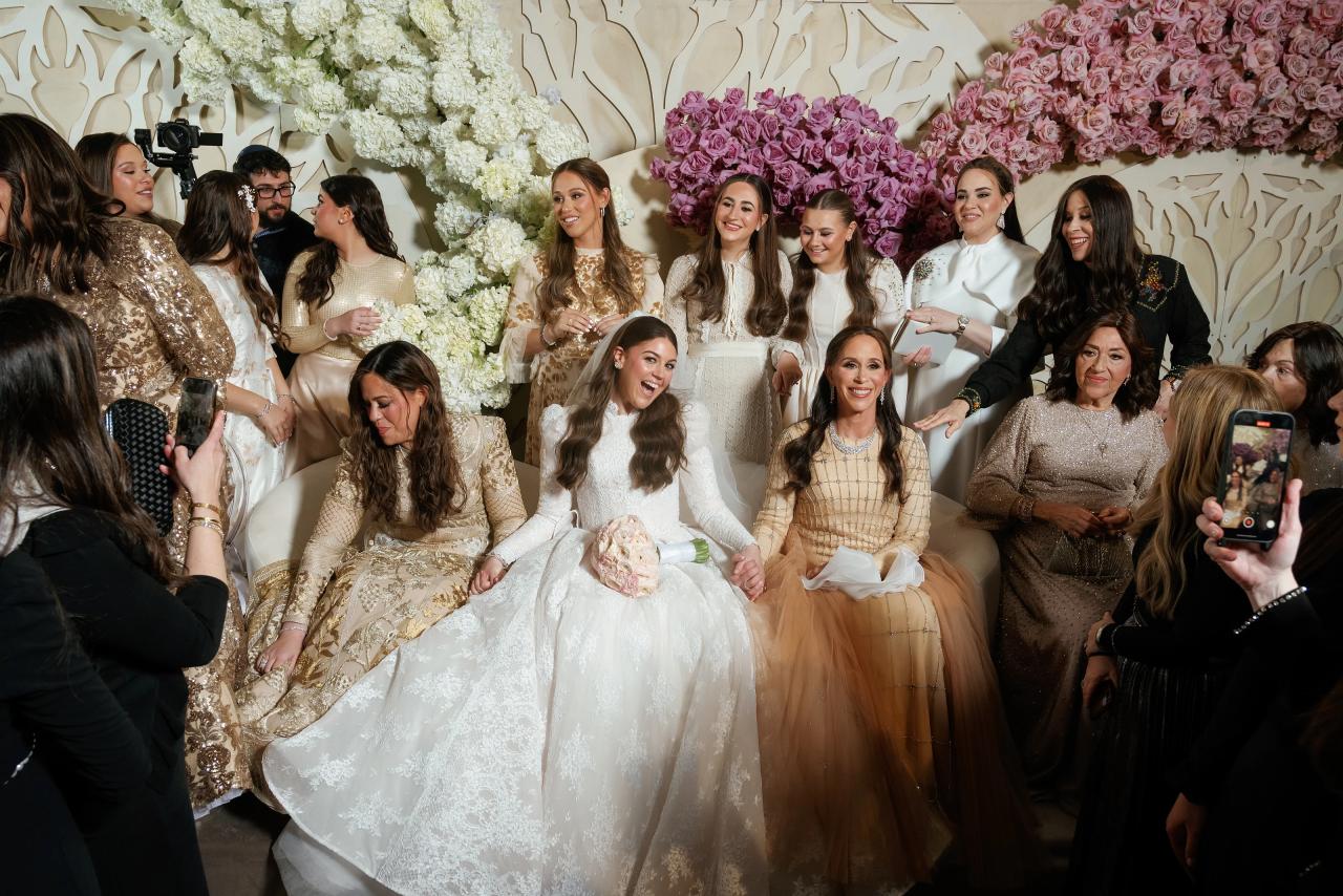 Bride seated at center with close family and guests around her, floral backdrop behind, guests clapping and one person holding up a phone at the edge of frame. Wedding photography at Lake Terrace, Lakewood, NJ.