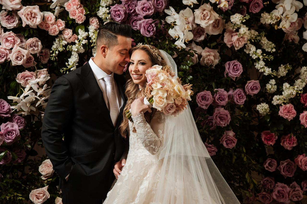 Bride and groom smiling closely together in front of a wall of pink, lilac and white flowers, with the bride holding a bouquet.