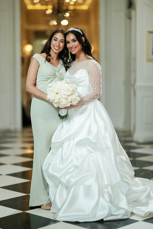 Two women in wedding attire stand close together smiling; one holds a white bouquet in a softly lit hallway with warm golden lights in the background.