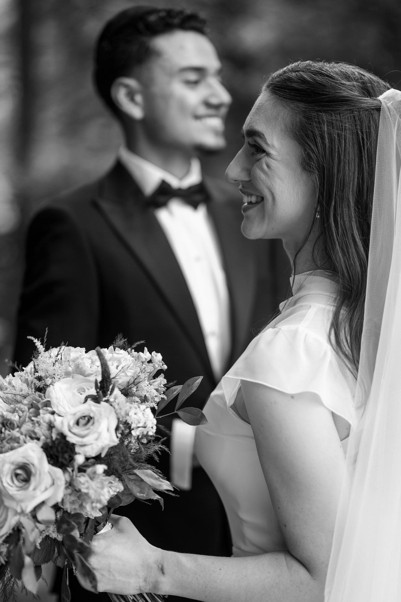 Black-and-white portrait of a smiling bride holding a bouquet, with the groom softly out of focus behind her.