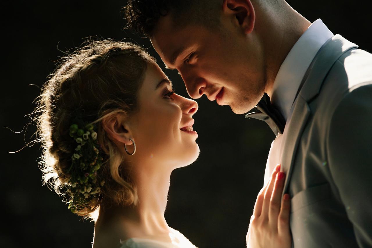 Intimate backlit wedding portrait of a bride and groom leaning face to face, with her hand resting on his light gray tuxedo.