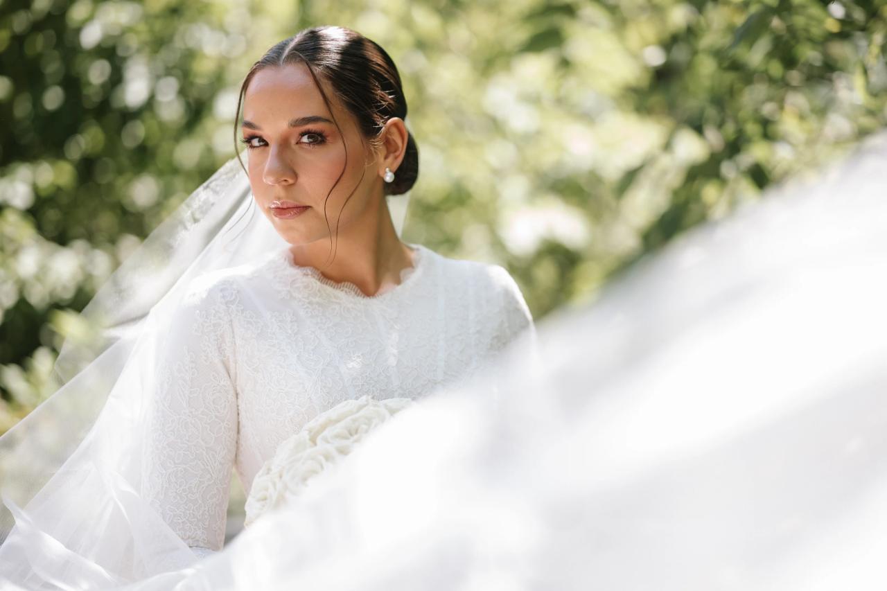 Close-up of the bride holding a white bouquet, framed by a softly blurred veil and sunlit greenery. From this Oholei Torah Ballroom wedding. Wedding photography.