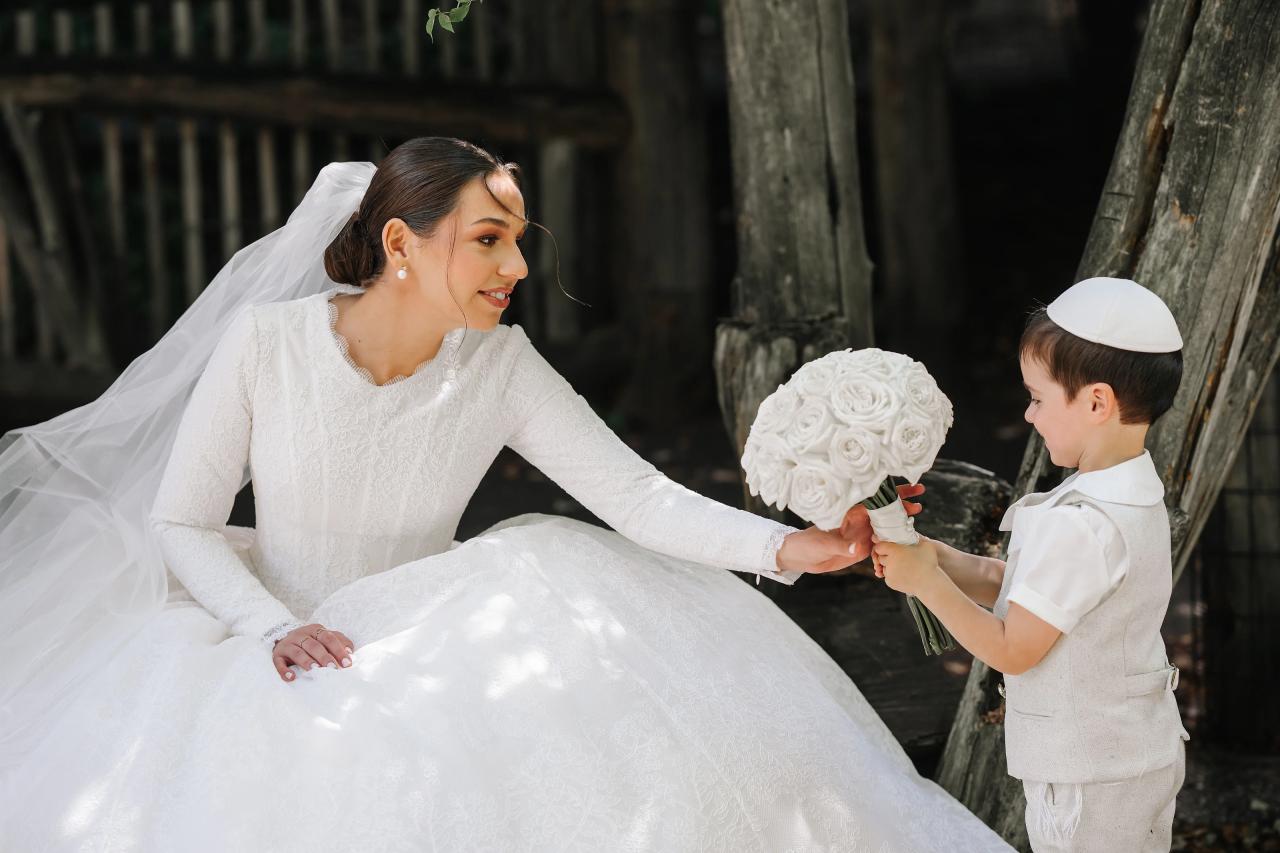 Bride smiling toward a small boy holding her white bouquet beside a tree. From this Oholei Torah Ballroom wedding. Wedding photography.