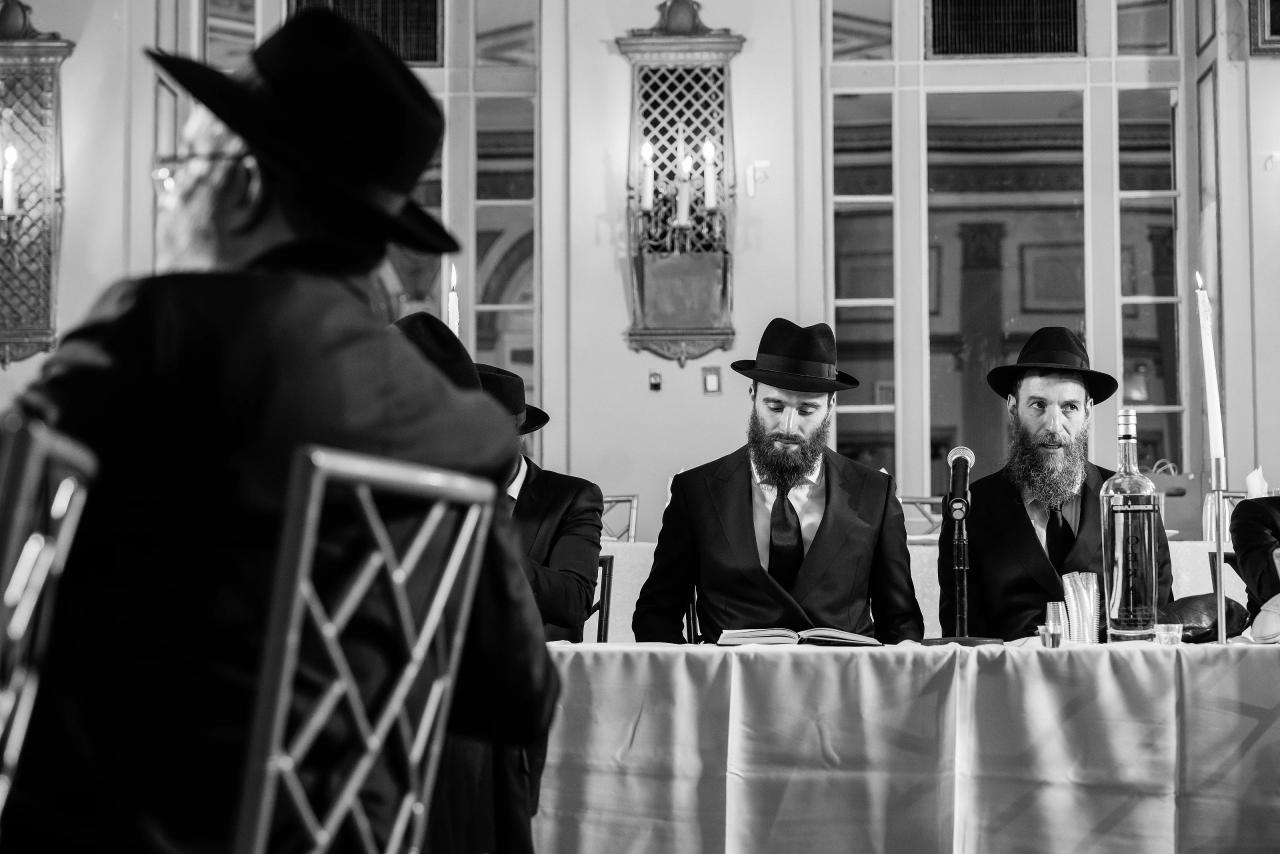Black-and-white view of the groom seated at the tisch, framed over a guest’s shoulder, with candles and mirrored walls behind him. From this Oholei Torah Ballroom wedding. Wedding photography.