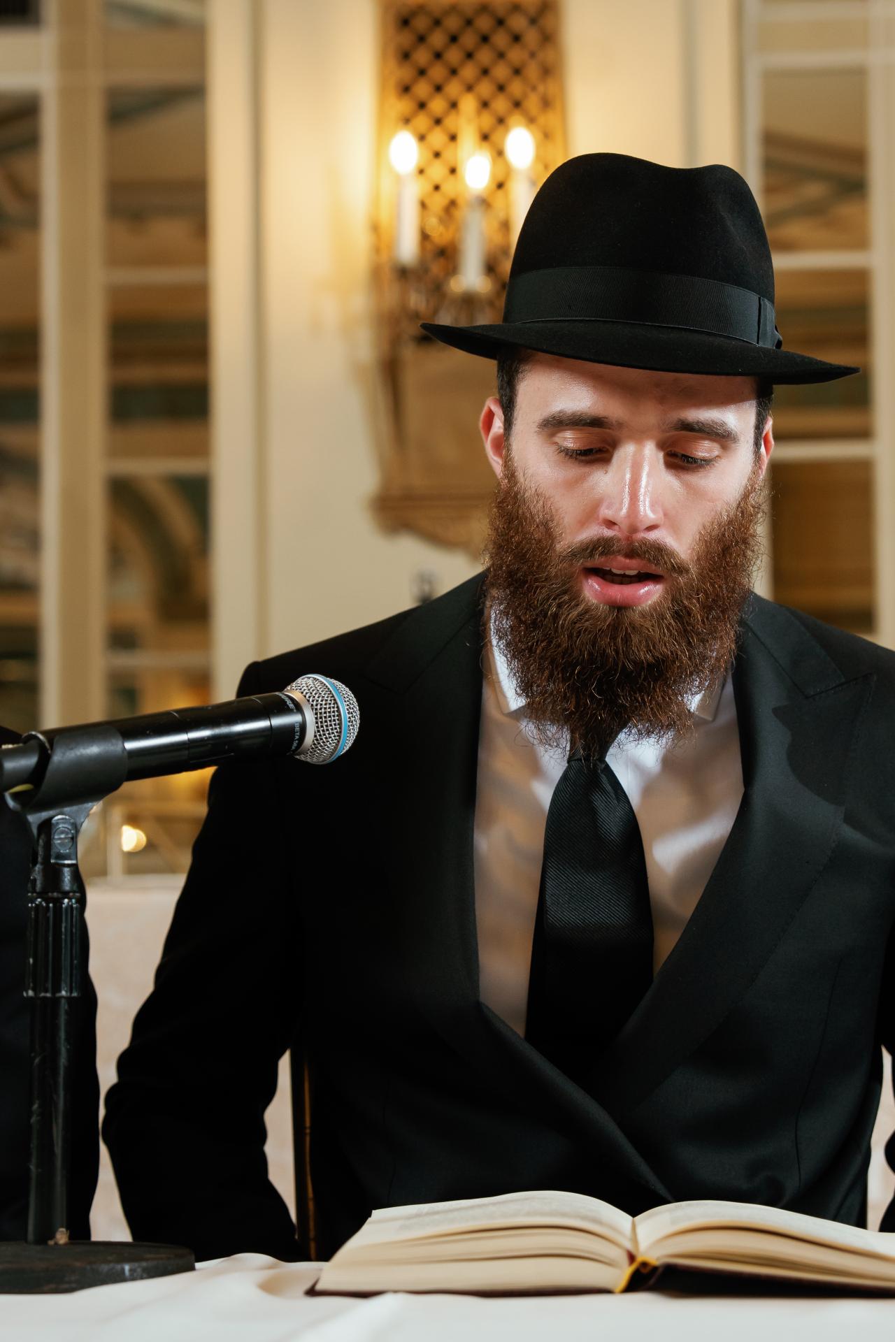 Groom reciting into a microphone beside an open book at the tisch. From this Oholei Torah Ballroom wedding. Wedding photography.