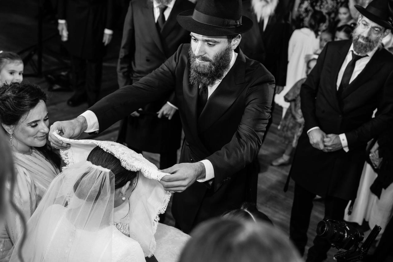 Black-and-white photograph of the groom lowering a lace veil over the bride during the bedeken, surrounded by family. From this Oholei Torah Ballroom wedding. Wedding photography.