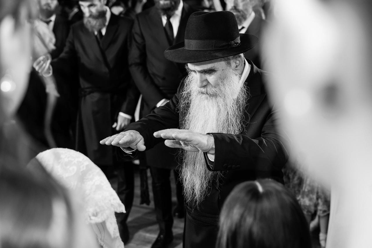 Black-and-white photograph of an elder holding his hands over the veiled bride in blessing, framed by guests in the foreground. From this Oholei Torah Ballroom wedding. Wedding photography.