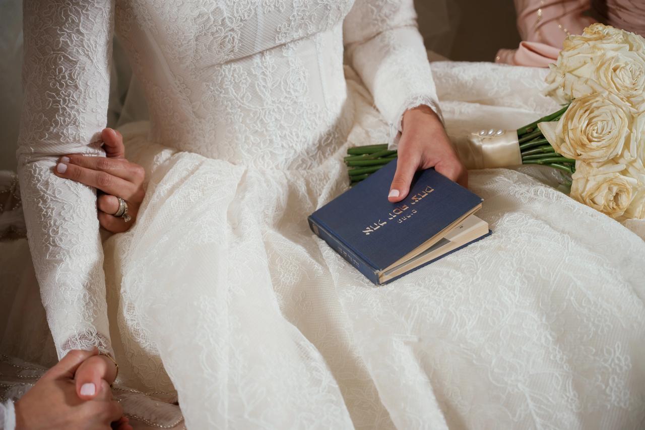 Close-up of hands resting on the bride’s lace gown beside a blue prayer book and white rose bouquet. From this Oholei Torah Ballroom wedding. Wedding photography.