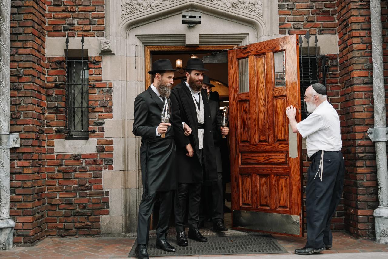 Groom stepping through a carved stone and wooden doorway in a red-brick building, accompanied by escorts carrying candles. From this Oholei Torah Ballroom wedding. Wedding photography.