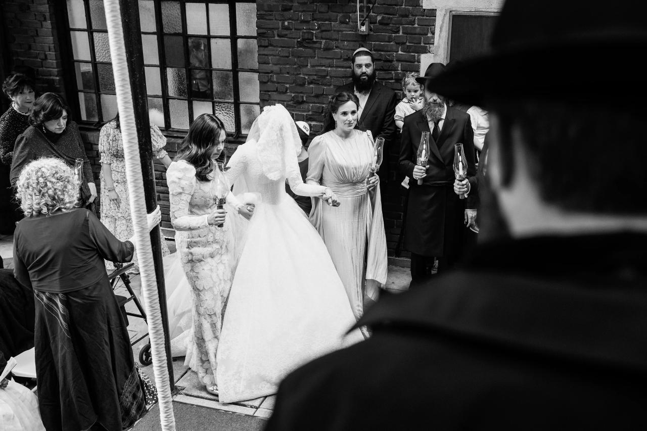 Black-and-white photograph of the veiled bride approaching the ceremony with candle-bearing escorts, seen over the groom’s shoulder. From this Oholei Torah Ballroom wedding. Wedding photography.