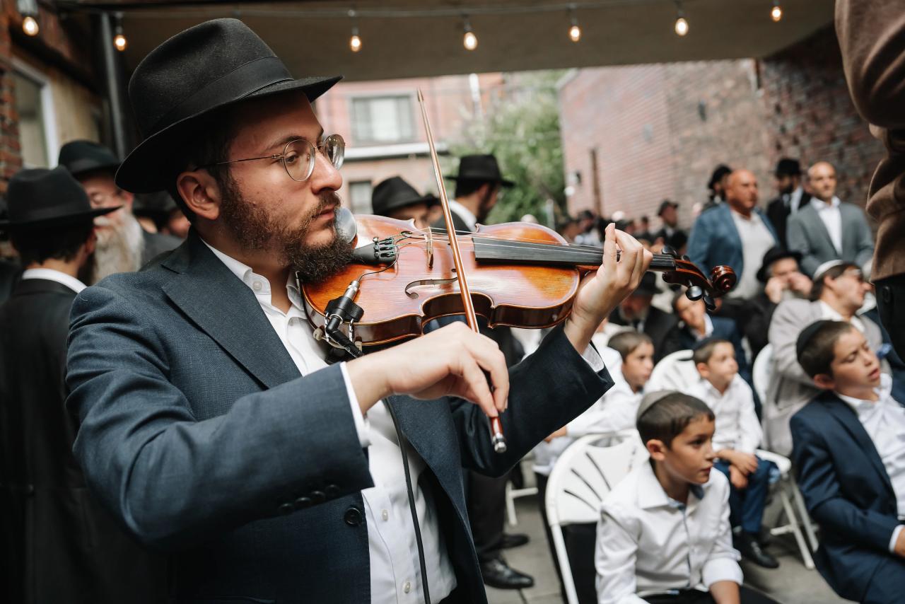 Violinist performing beside wedding guests in a brick-walled courtyard beneath strings of lights. From this Oholei Torah Ballroom wedding. Wedding photography.