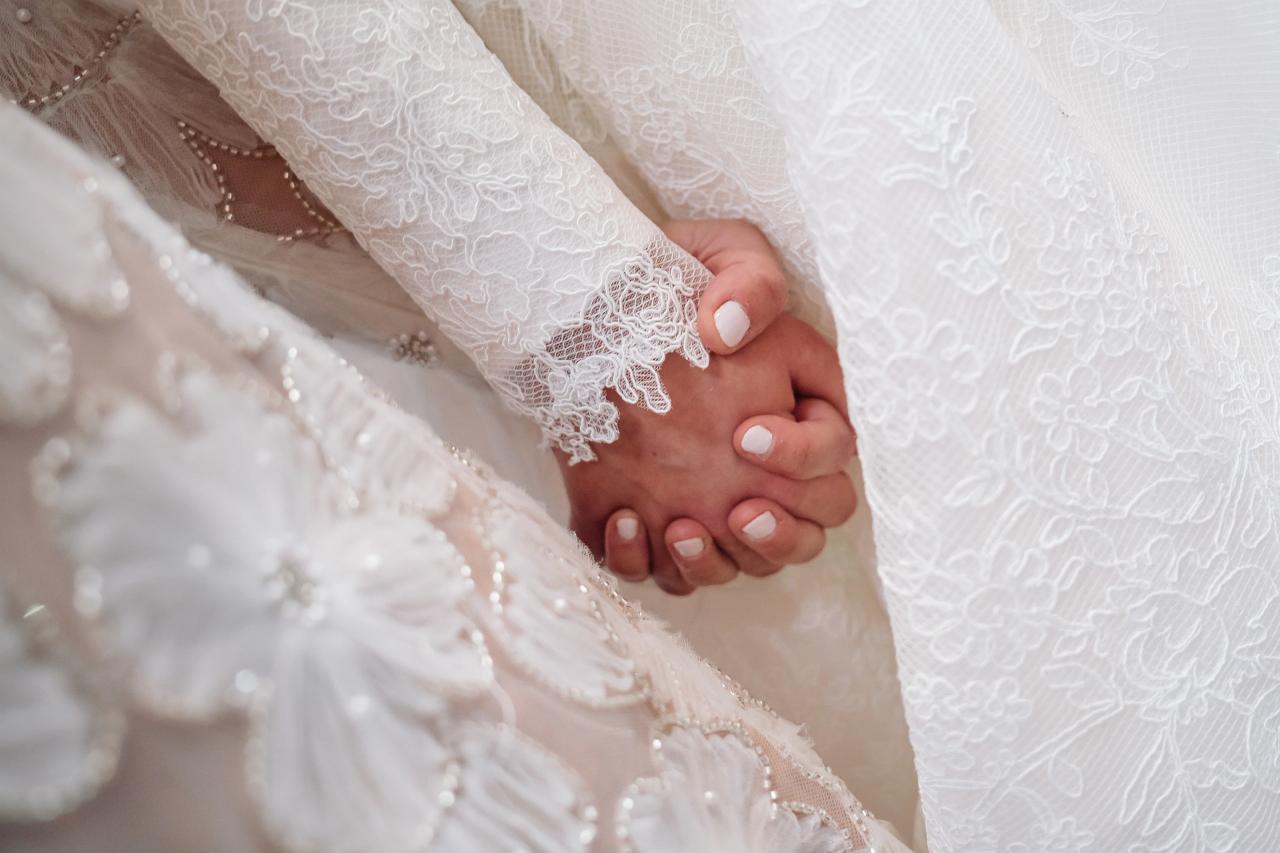 Close-up of the bride's clasped hands, lace sleeves and beaded gown beneath a patterned veil. From this Oholei Torah Ballroom wedding. Wedding photography.